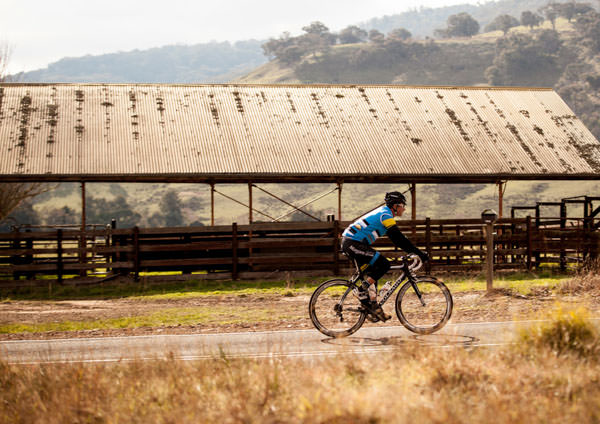A man cycling past old wood and tin farm buildings