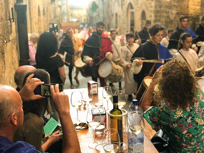 People have a glass of wine in a Tuscan street as a colourful festival passes
