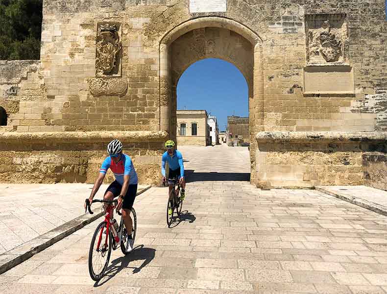 Two rider cycling through an old stone arch in a Puglian town