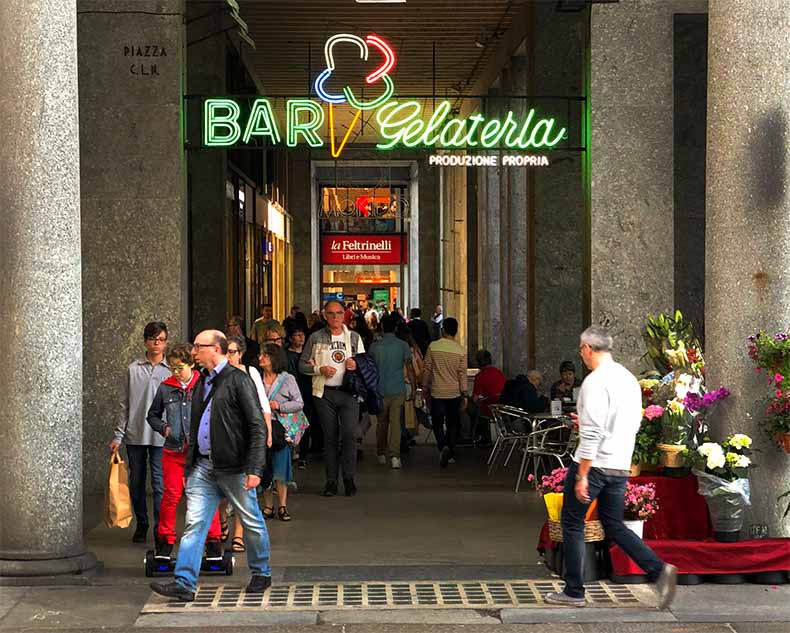 People walking under a portico and neon gelati sign