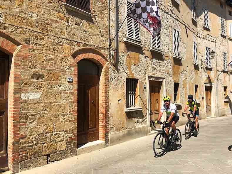 Two riders cycling through a stone Tuscan town