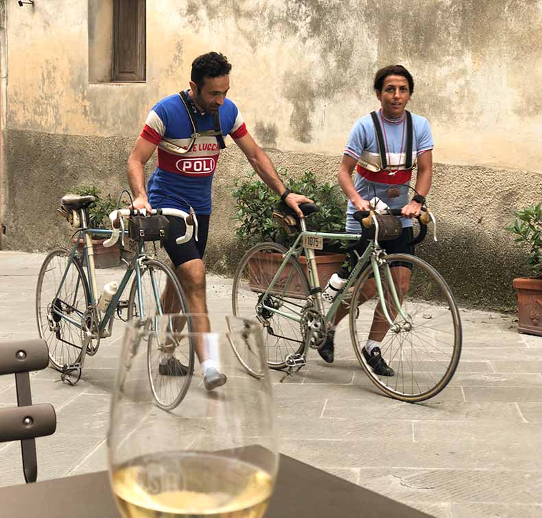 Two people wheeling their steel bikes past a bar in Italy